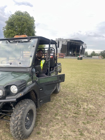 Rasheed in hi-vis at the wheel of a site buggy on a festival field, with the main stage behind him